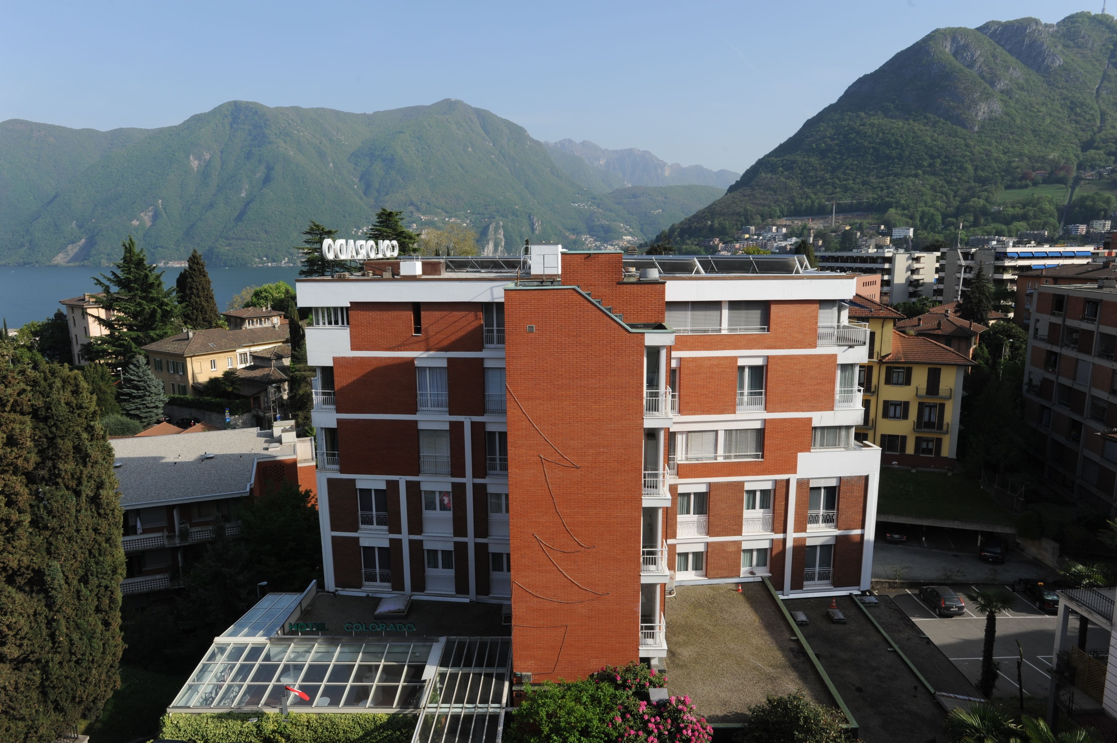 Un immeuble à plusieurs étages doté d’une terrasse sur le toit, entouré d’autres bâtiments et dominant un paysage montagneux.