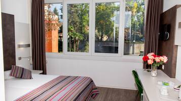 Habitación de hotel con cama, escritorio, silla, televisor y ventana con cortinas.