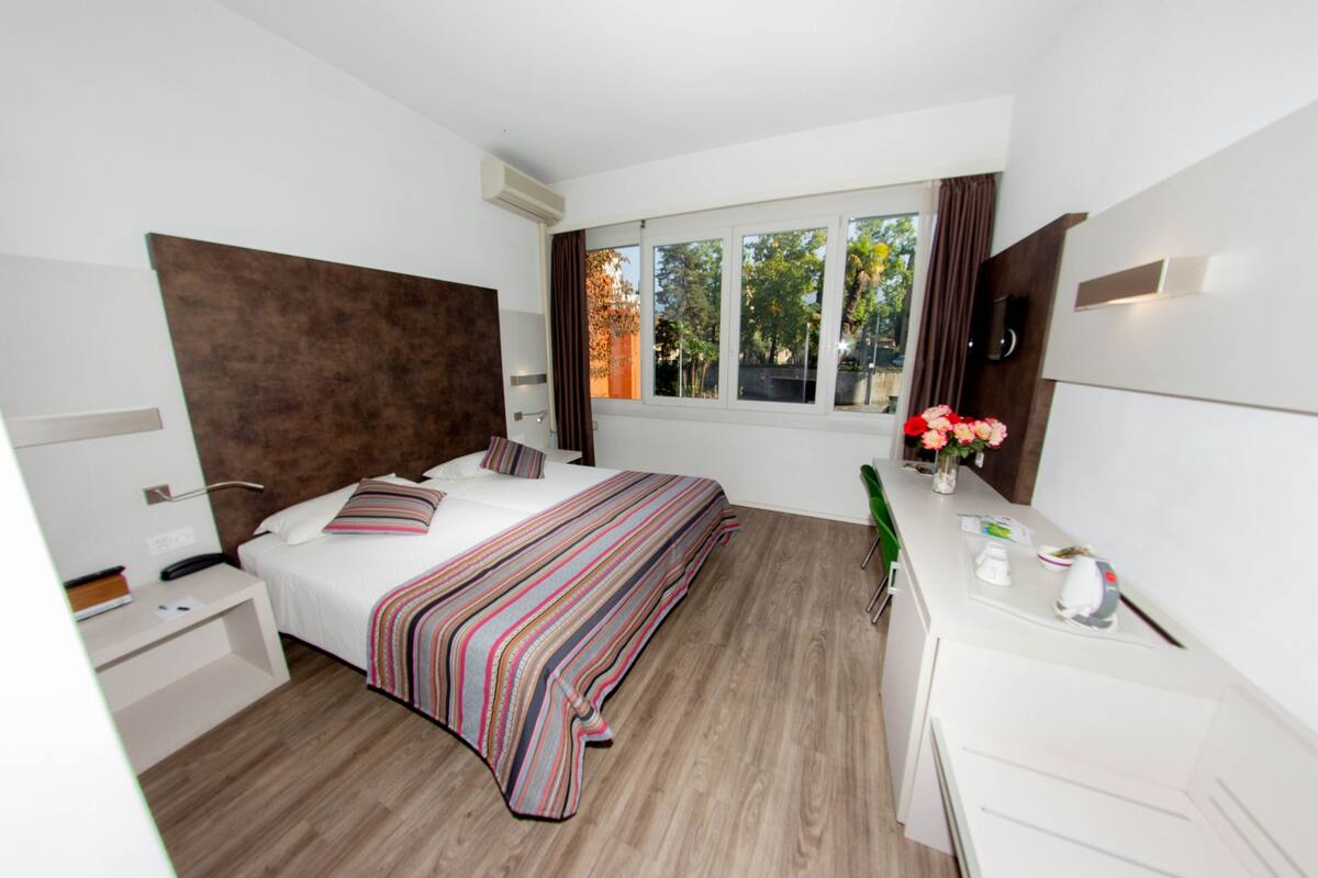 Ett modernt hotellrum med en stor säng, ett skrivbord, en stol, ett fönster med gardiner och en väggmonterad TV.