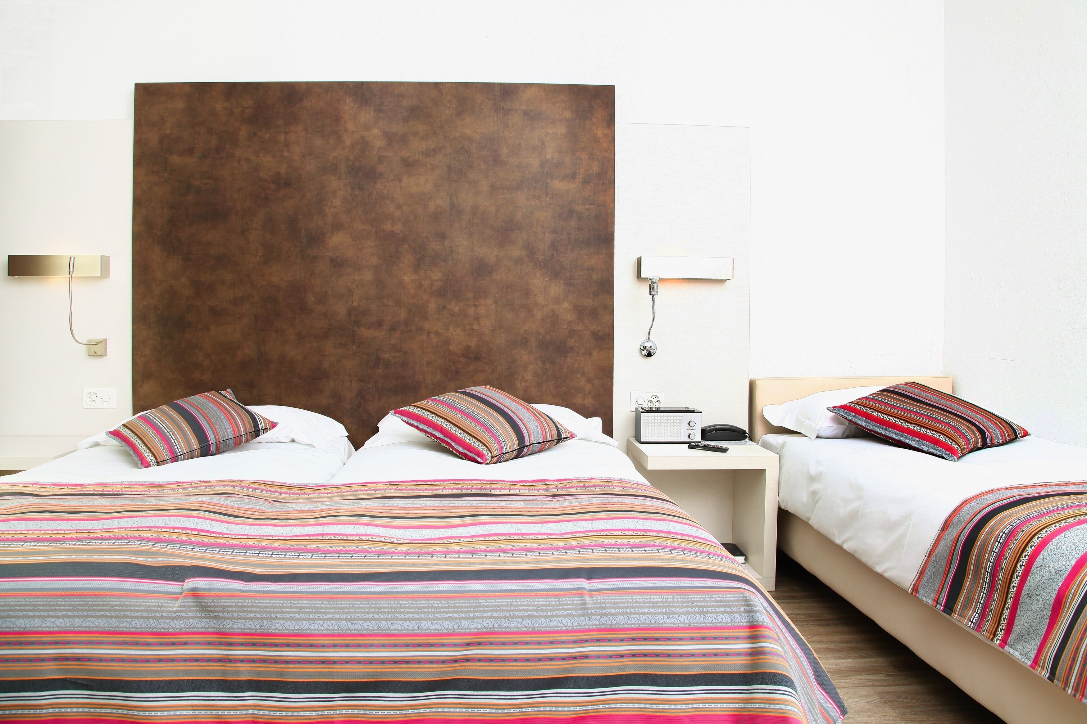 A hotel room with a large bed, a smaller bed, a wooden headboard, a bedside table with a lamp, and a wall-mounted light fixture.