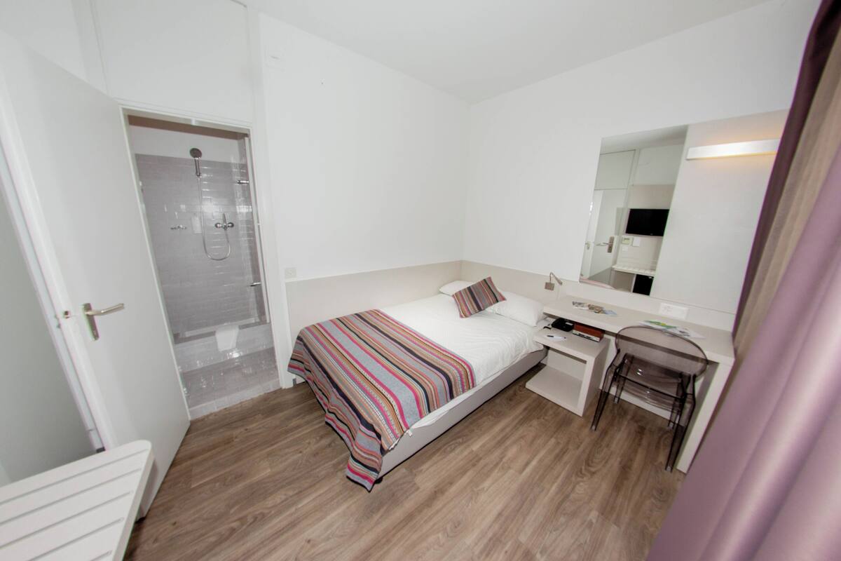 Ett hotellrum med en säng, ett nattduksbord, ett skrivbord, en stol och en dusch.