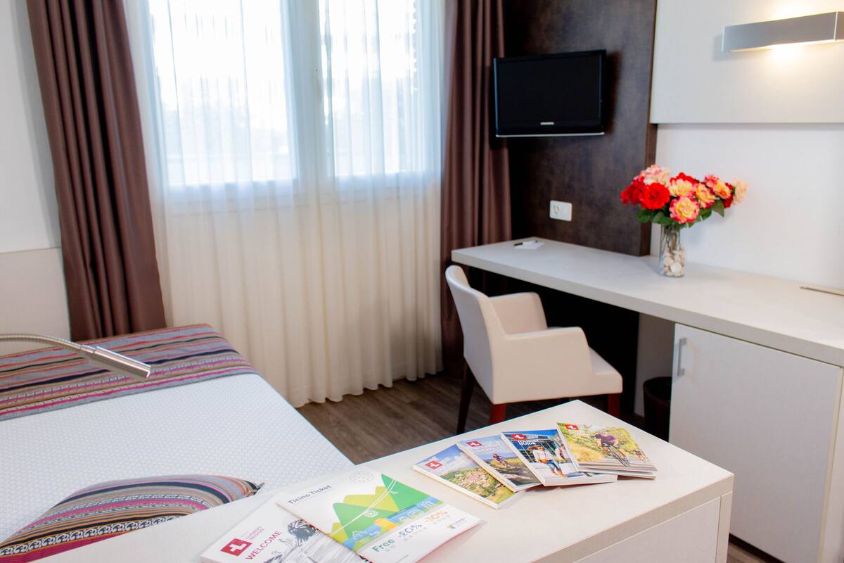 Ett hotellrum med en säng, ett skrivbord med en stol, en tv, ett litet kylskåp och en vas med blommor.
