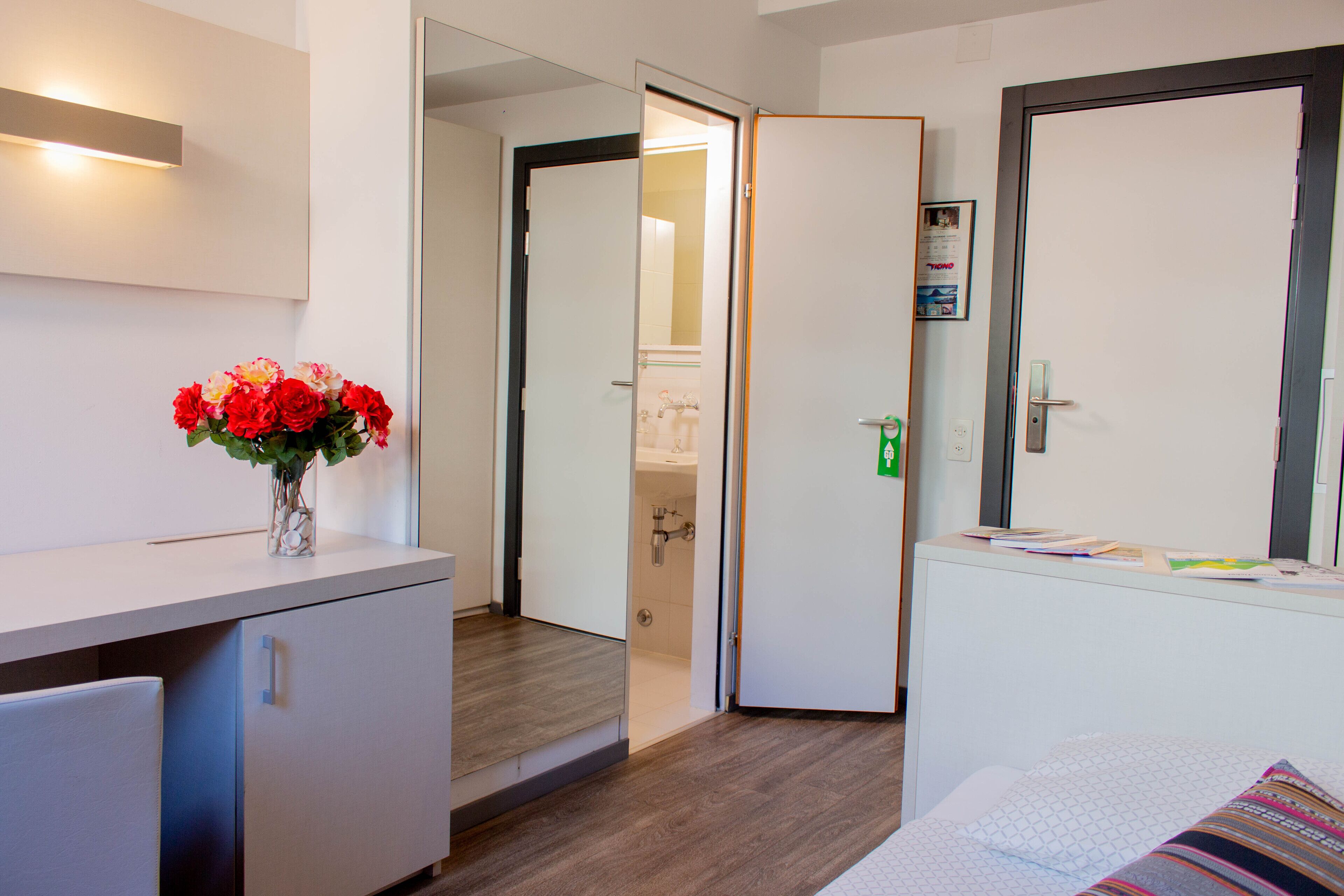 Habitación de hotel con piso de madera, un escritorio con un florero y una cama con manta a rayas, y una puerta que da a un baño.