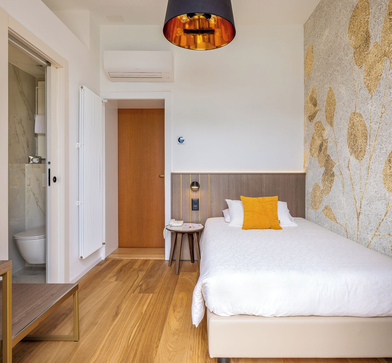 Een moderne hotelkamer met een houten vloer, een bed met wit beddengoed en een geel kussen, een klein rond tafeltje met een telefoon, en een wandgemonteerde airconditioning.