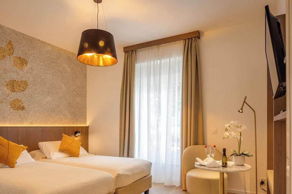 A hotel room with two beds, a large window with curtains, a desk with a chair, a small table with a lamp, and a wall-mounted TV.