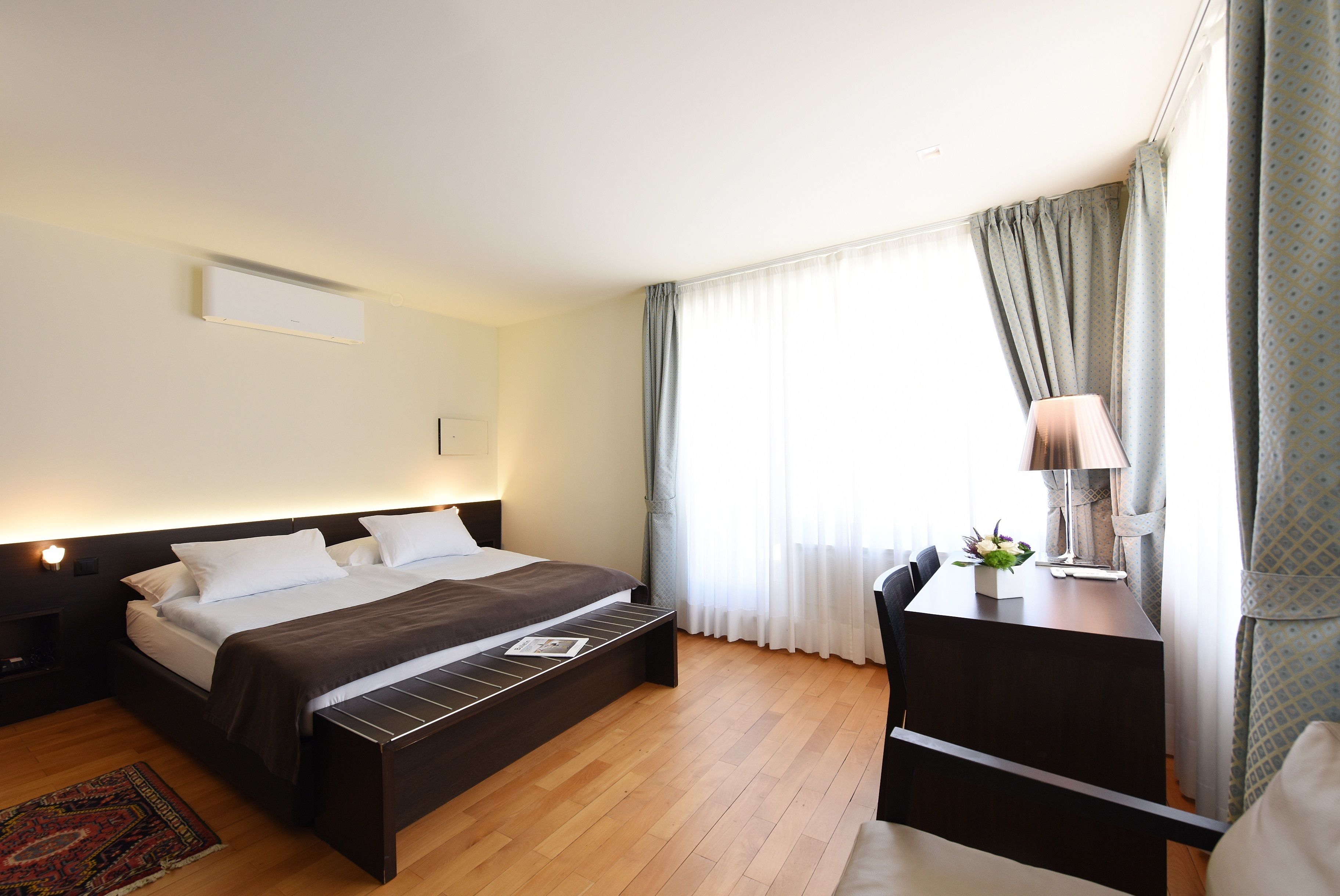 A modern hotel room with a large bed, bedside table, desk, and a view of the outdoors through sheer curtains.