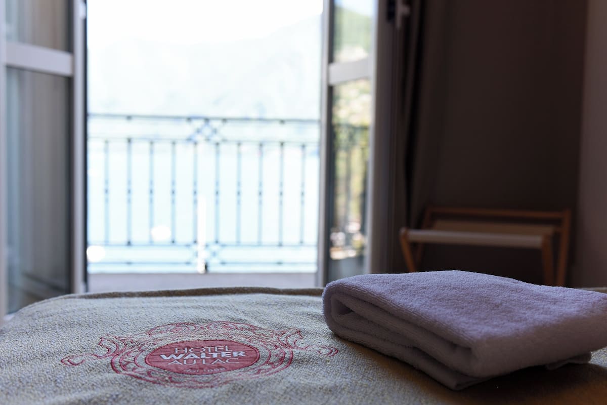 A neatly folded towel on a bed with a towel with a logo, a window with a view of a balcony, and a chair in the corner.