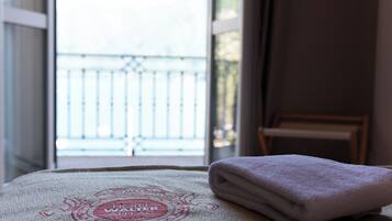 Ein ordentlich gefaltetes Handtuch auf einem Bett, ein Handtuch mit einem Logo, ein Fenster mit Blick auf einen Balkon und ein Stuhl in der Ecke.