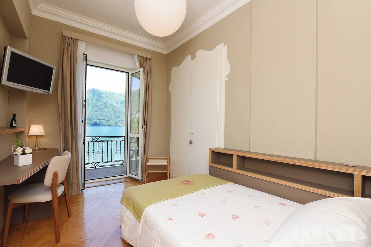 A hotel room with a bed, a desk with a chair, a television, and a view of a balcony with a scenic landscape.