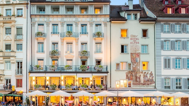 A hotel with a prominent sign, multiple balconies with plants, and a reflection in the water. - Des Alpes Luzern (Lucerne)