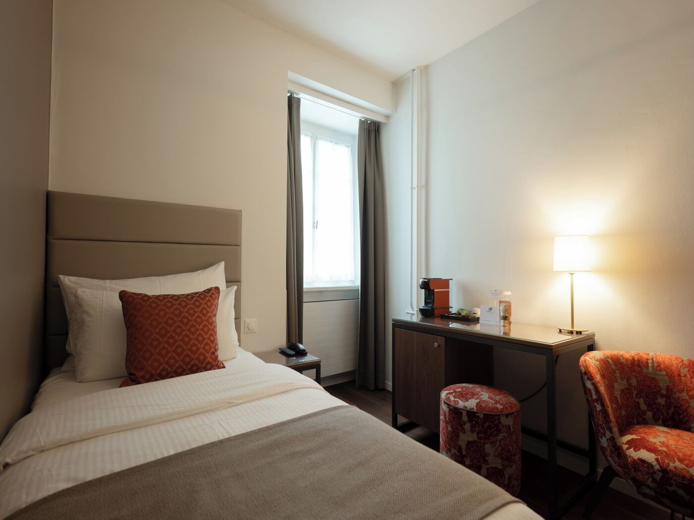 A hotel room with a bed, a desk, a chair, a lamp, and a window with curtains.