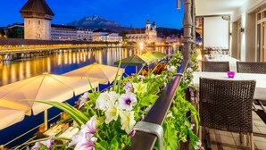A riverside terrace with outdoor seating, a view of a historic building, and a tower in the background. - Des Alpes Luzern (Lucerne)
