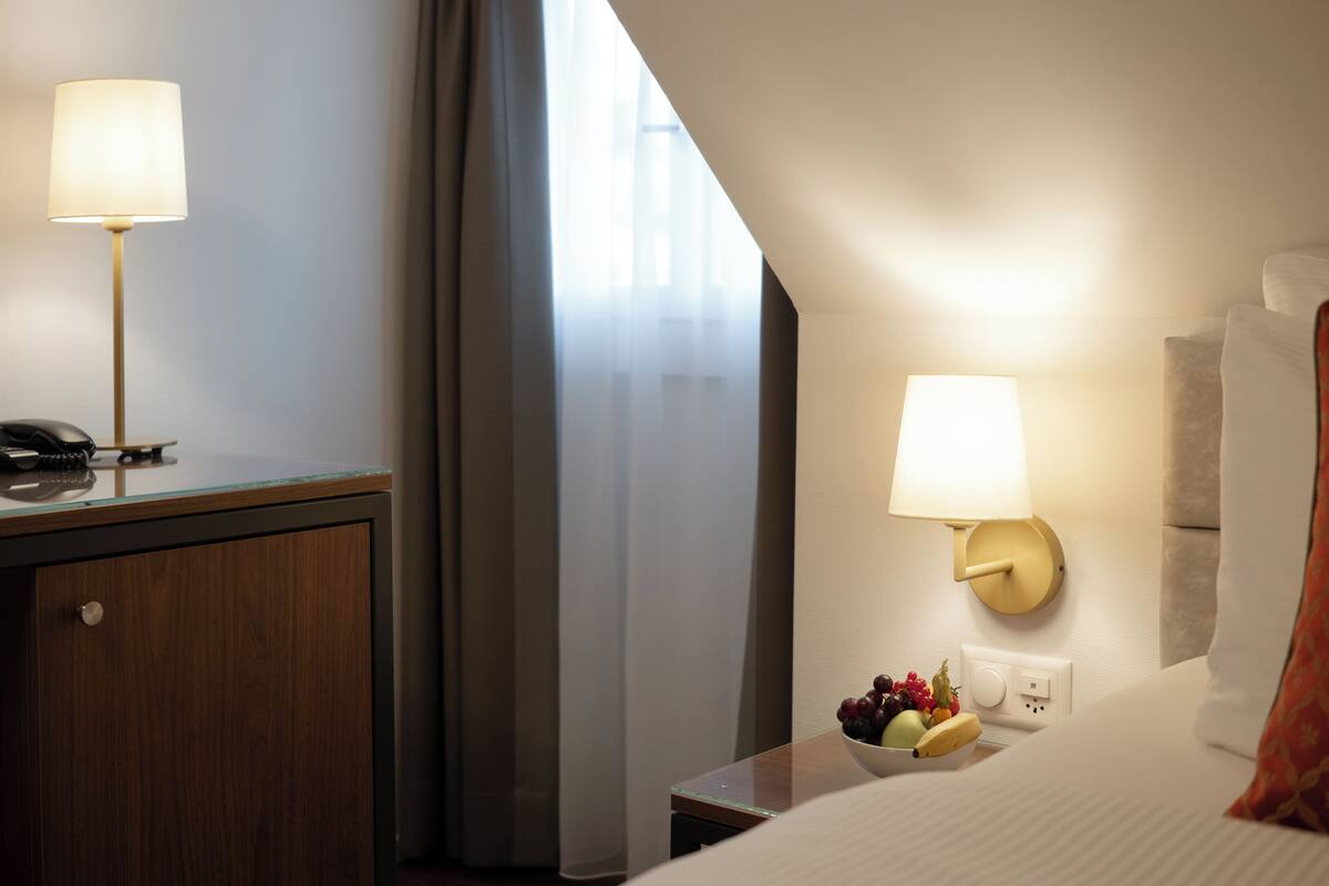 Quarto de hotel com escrivaninha, duas luminárias, mesa de cabeceira com frutas e uma cama com travesseiro.
