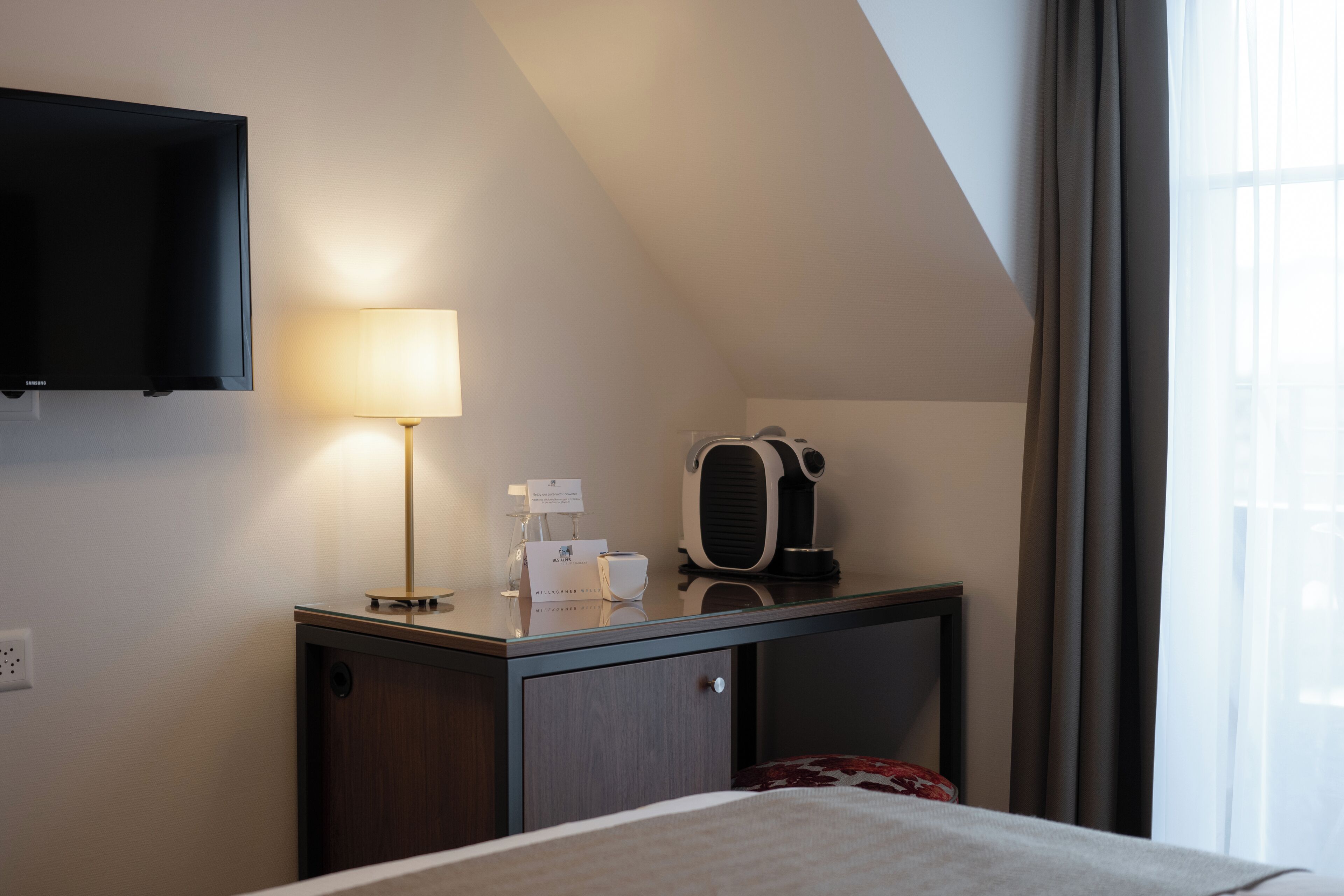 A hotel room with a bed, a bedside table with a lamp, a coffee maker, and a TV mounted on the wall.