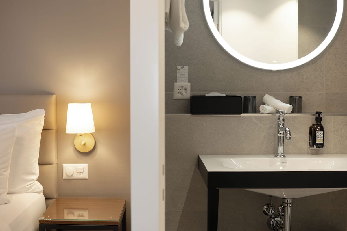 Quarto de hotel com cama, mesa de cabeceira com abajur, espelho fixado na parede e banheiro com pia e artigos de higiene pessoal.
