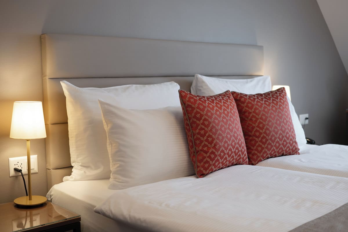 Cama bem arrumada com almofadas brancas e estampadas, um abajur e uma tomada na parede.