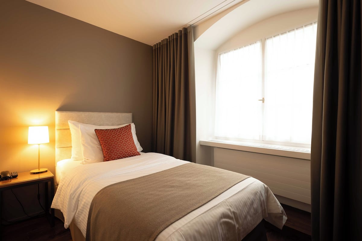 Quarto de hotel com cama, luminária de cabeceira, janela com cortinas e mesa de cabeceira com telefone.
