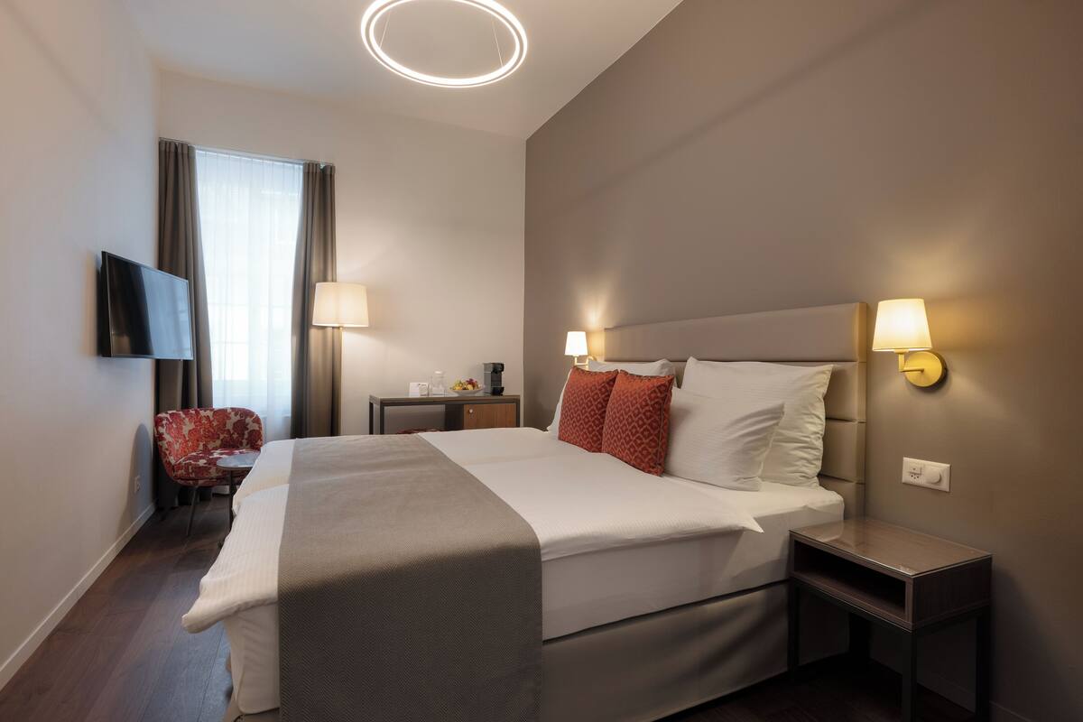 Quarto de hotel moderno com uma cama grande, duas mesinhas de cabeceira com abajures, uma televisão de tela plana na parede, uma escrivaninha pequena com cadeira e uma janela com cortinas.