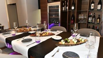 A dining area with multiple tables set for a meal, each with plates, cutlery, and wine glasses. A wine rack with bottles is visible in the background.