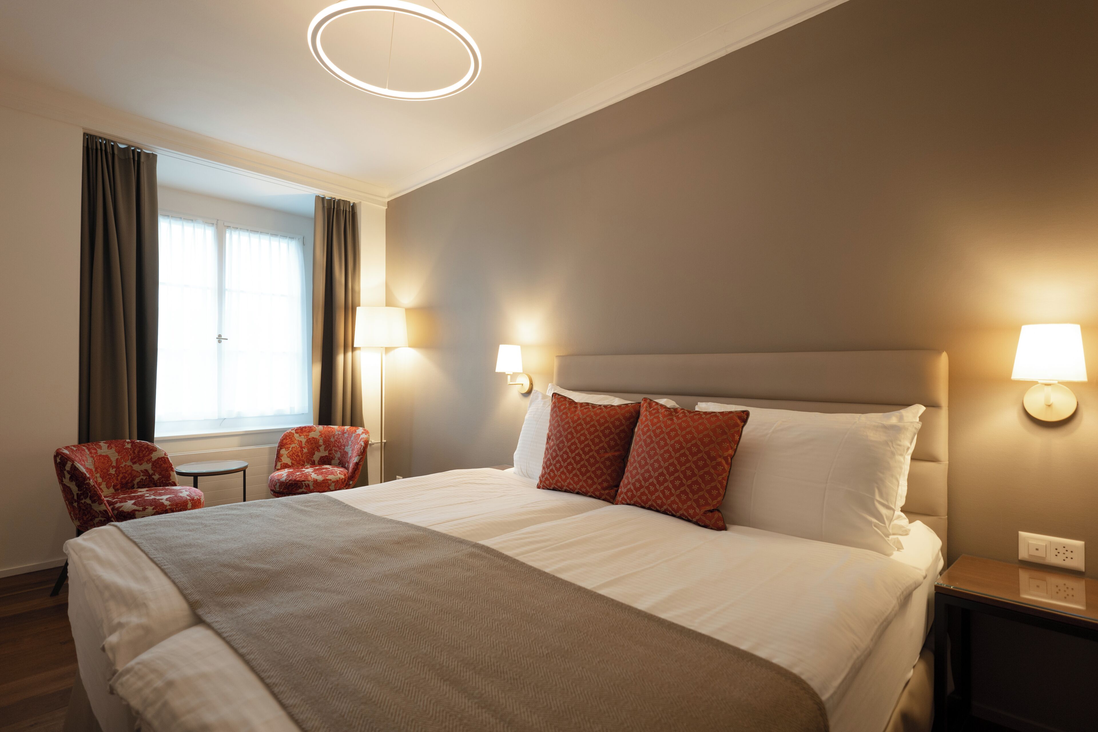A hotel room with a large bed, two chairs, a nightstand, and a window with curtains.