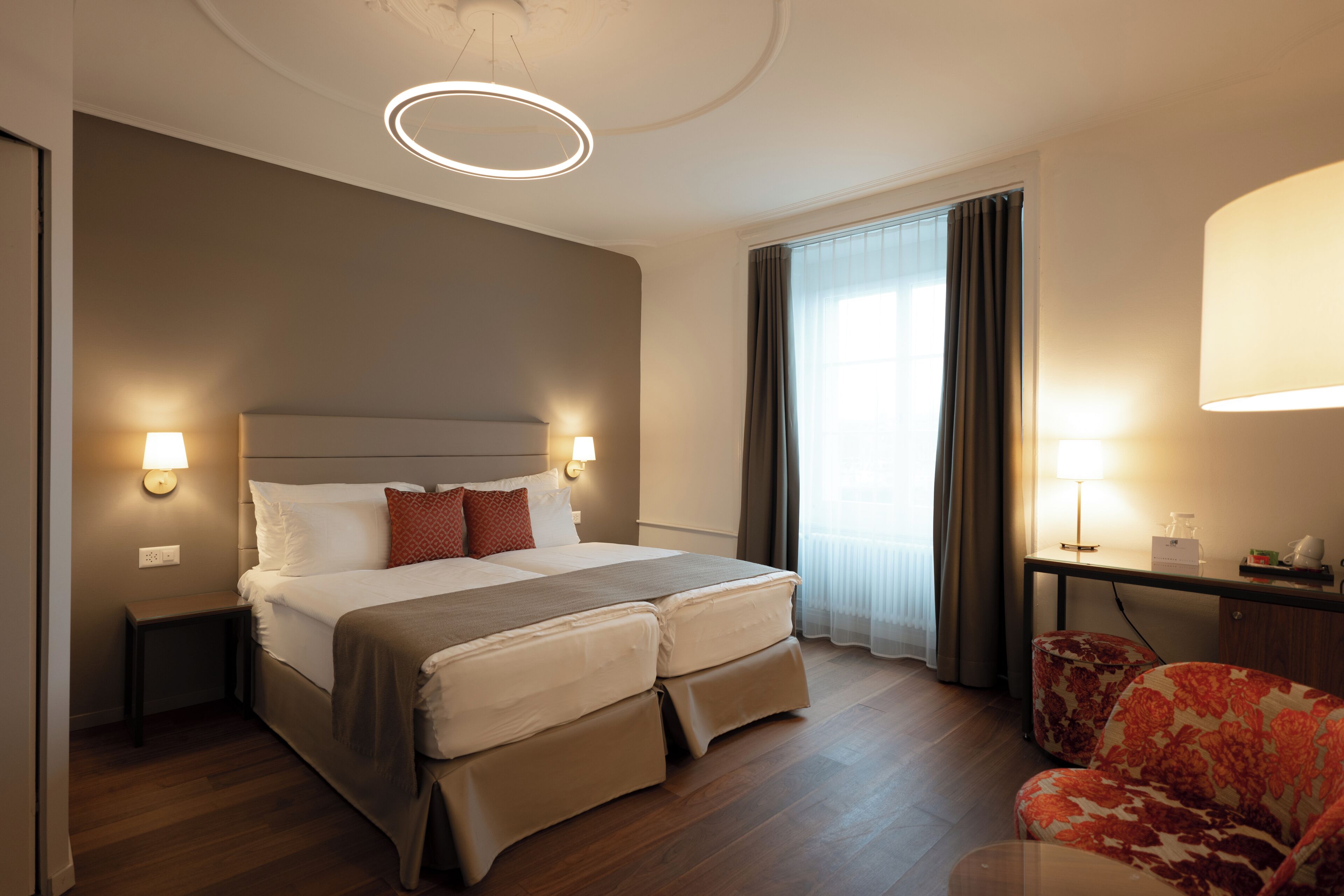 A hotel room with a bed, bedside lamps, a desk with a lamp, a chair, and a large window with curtains.