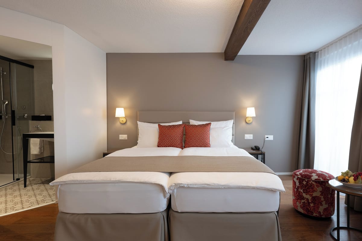 Uma cama de casal com duas almofadas, uma mesinha de cabeceira com abajur e uma pequena mesa redonda com frutas.