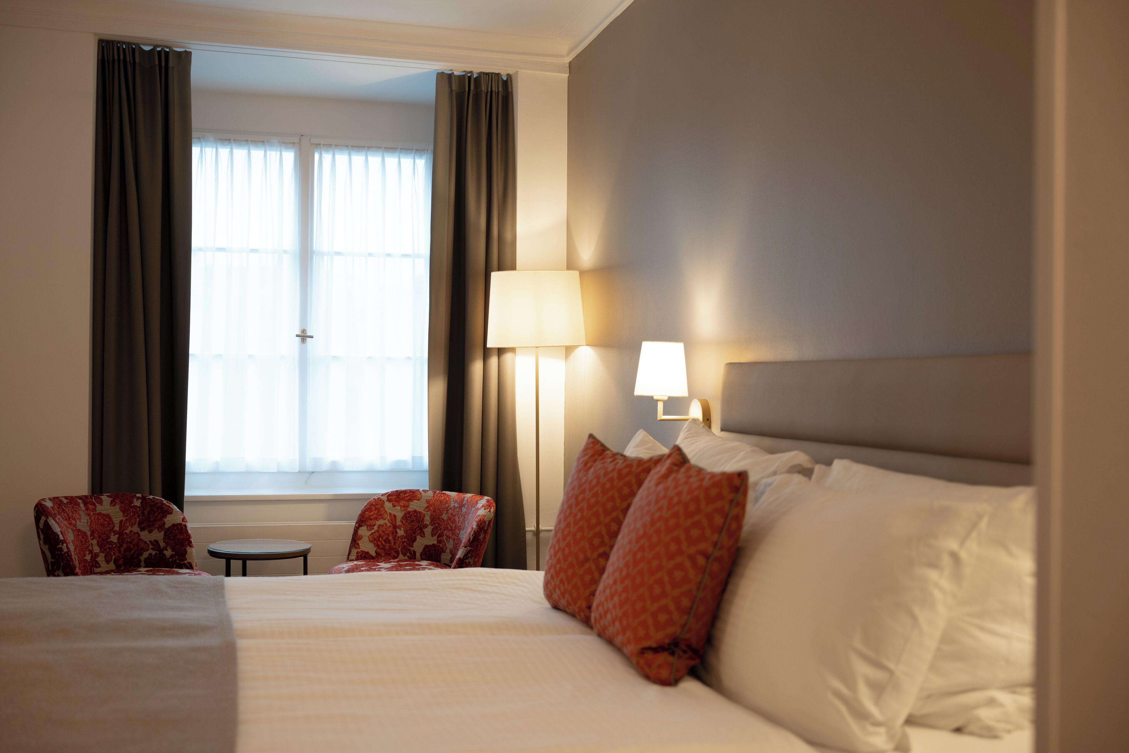 A hotel room with a bed, two chairs, a lamp, and a window with curtains.