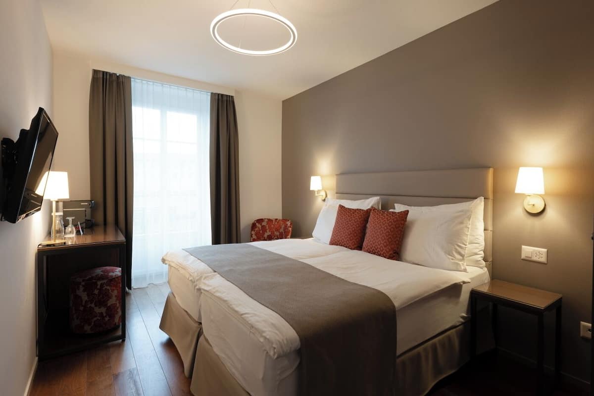 Quarto de hotel com cama grande, abajures, televisão de tela plana, mesinha e janela com cortinas.