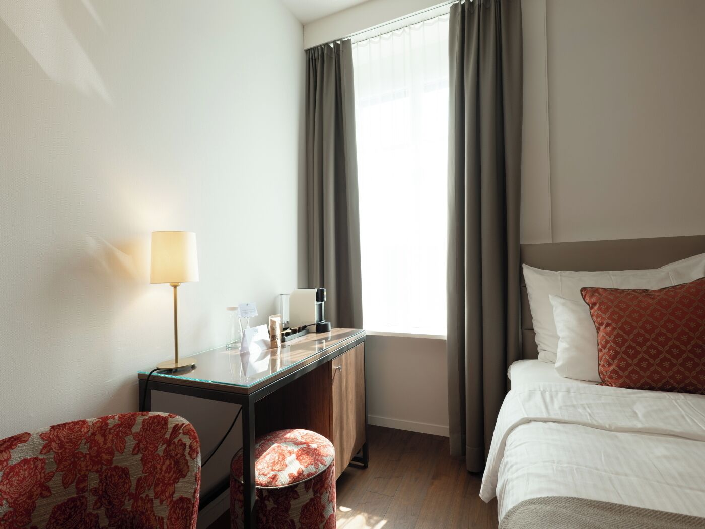 A hotel room with a bed, a desk with a lamp, a chair, and a window with curtains.