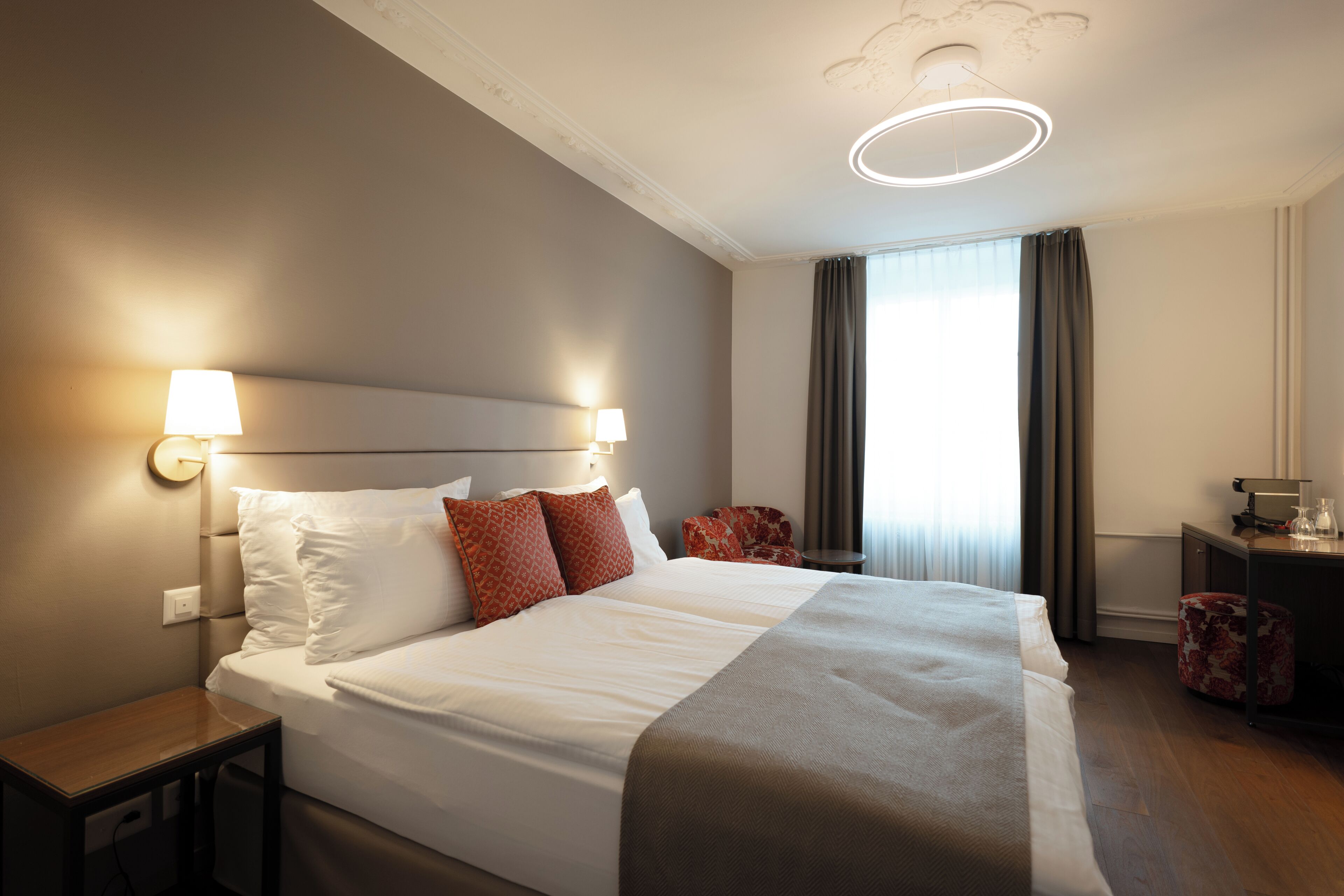 Ein Hotelzimmer mit einem großen Bett, zwei Nachttischlampen, einem Schreibtisch mit Lampe, einem Sessel und einem Fenster mit Vorhängen.