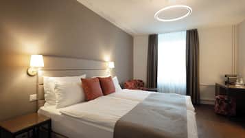 Ein Hotelzimmer mit einem großen Bett, zwei Nachttischlampen, einem Schreibtisch mit Lampe, einem Sessel und einem Fenster mit Vorhängen.