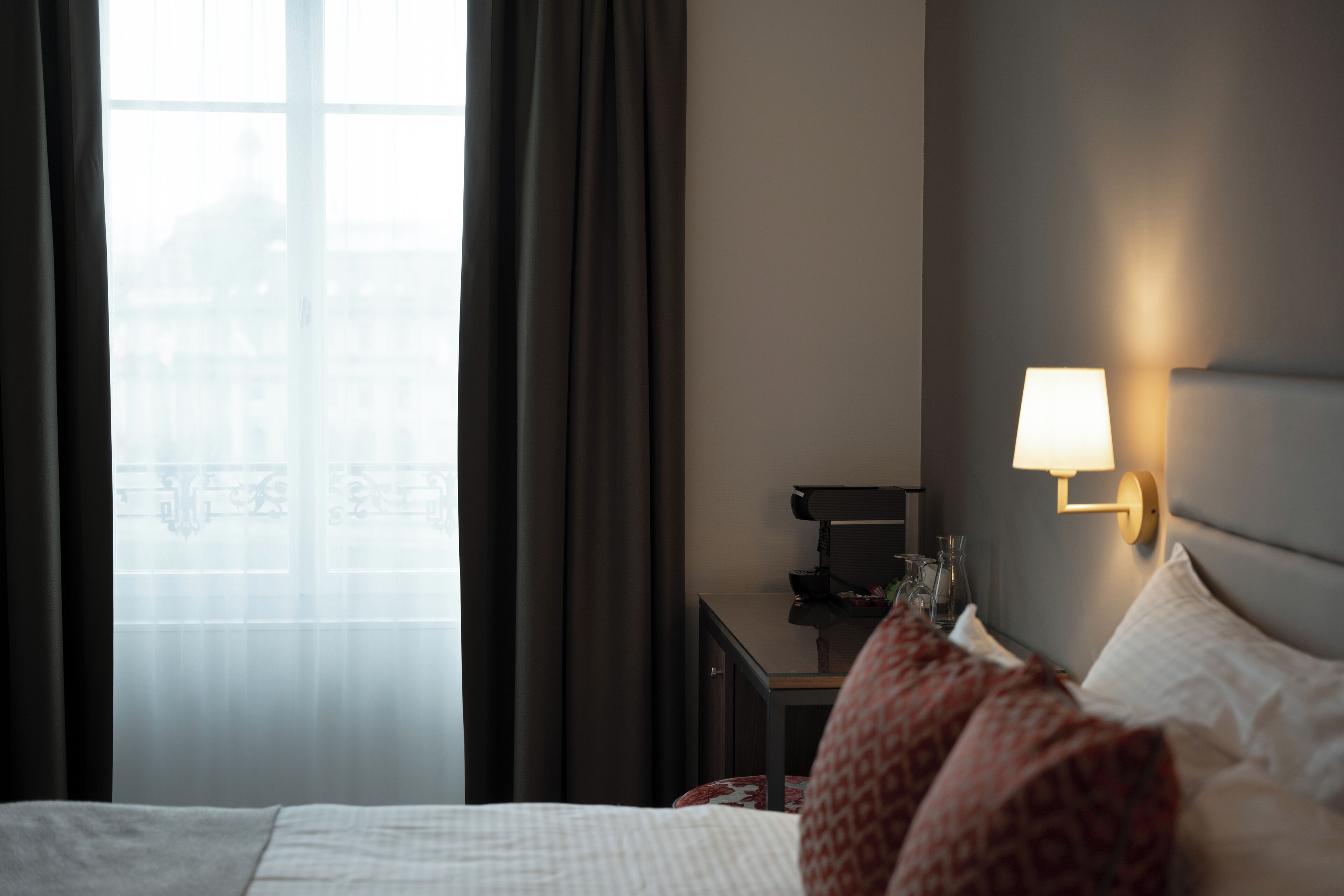 Een hotelkamer met een bed, een nachtkastje, een lamp en uitzicht op een gebouw door een raam.