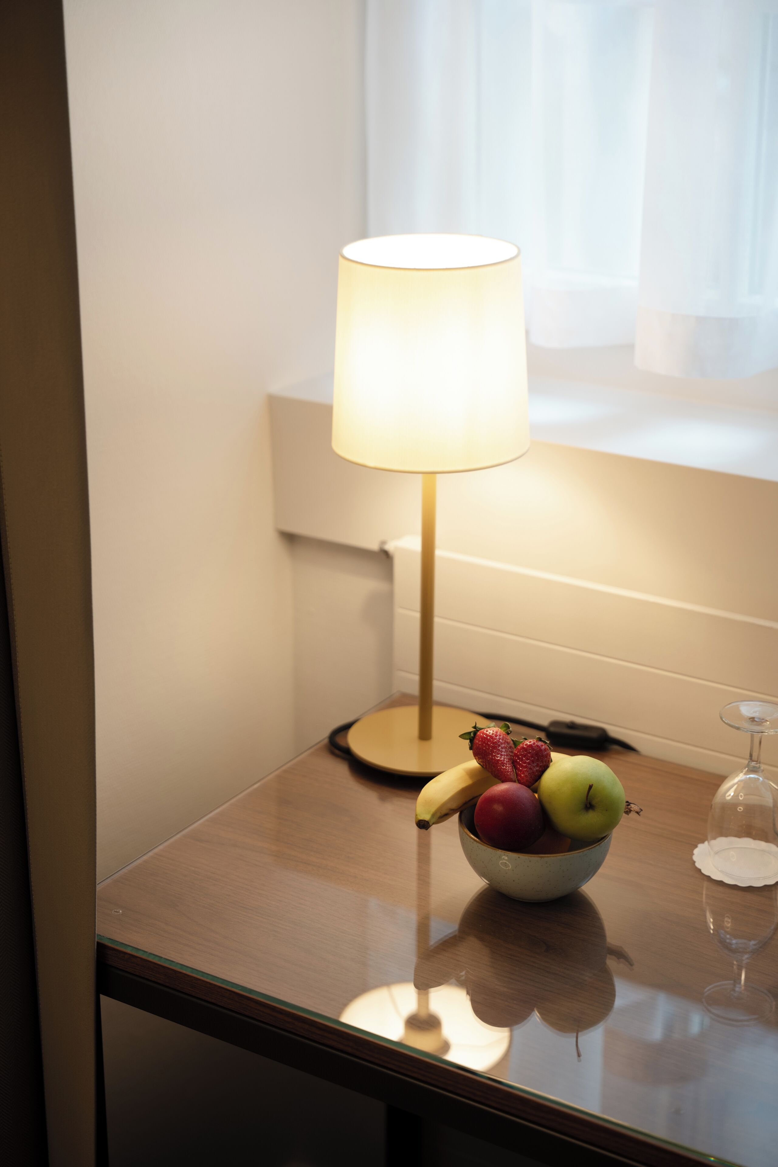 Una mesa con un bol de fruta, una lámpara y una copa de vino.