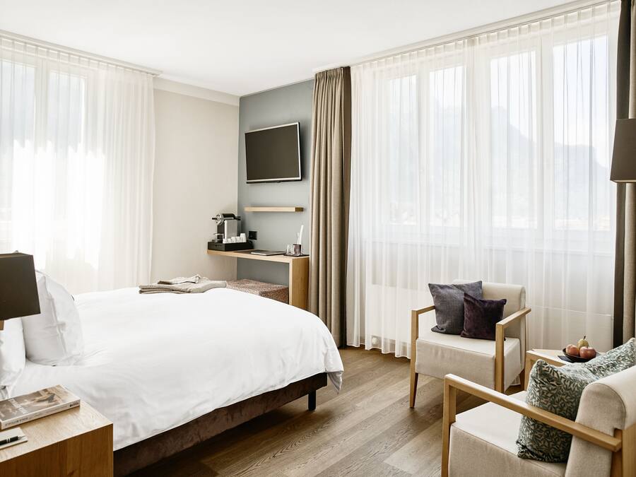 A modern hotel room with a large bed, a desk with a coffee maker, a TV mounted on the wall, and a seating area with a chair and cushions.