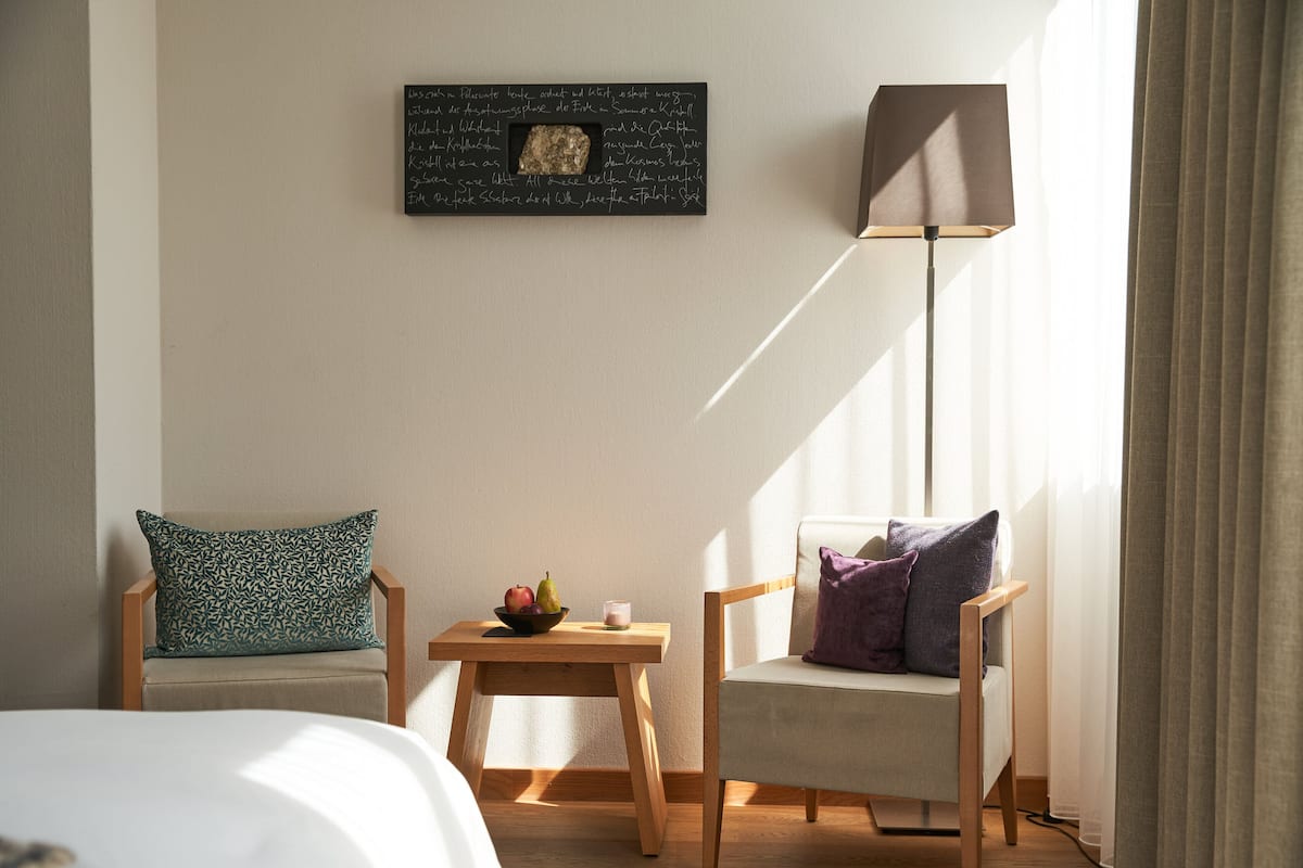 Ein Hotelzimmer mit einem Bett, zwei Stühlen, einem kleinen Tisch mit einer Obstschale, einer Kerze und einem Wandbild.