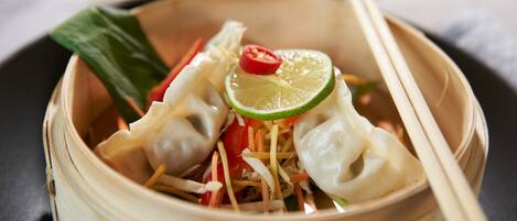 Una vaporera de bambú con comida, incluyendo dumplings y verduras, además de palillos sobre un plato.