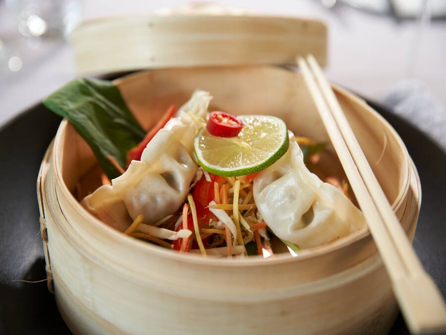 A bamboo steamer with food, including dumplings and vegetables, and chopsticks on a plate.