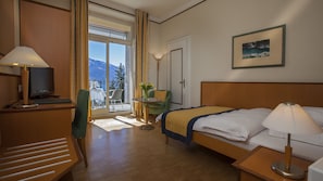 A hotel room with a bed, a desk with a computer, a chair, a small table with a vase of flowers, and a view of mountains through the window. - Esplanade Hotel Resort & Spa (Minusio)