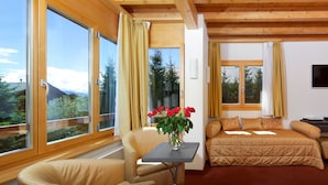 A room with a bed, a chair, a table with a vase of flowers, and large windows with curtains. - La Prairie (Crans-Montana)