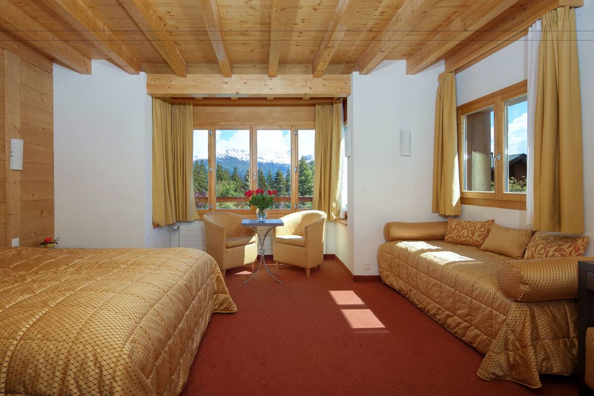 Une chambre avec deux lits, une table et une chaise, offrant une vue sur les montagnes grâce à de grandes fenêtres.