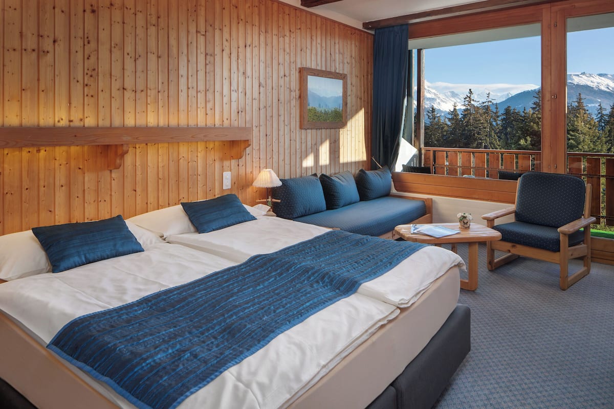 Une chambre avec deux lits, un canapé, une chaise et une table. Il y a une grande fenêtre qui offre une vue sur les montagnes.