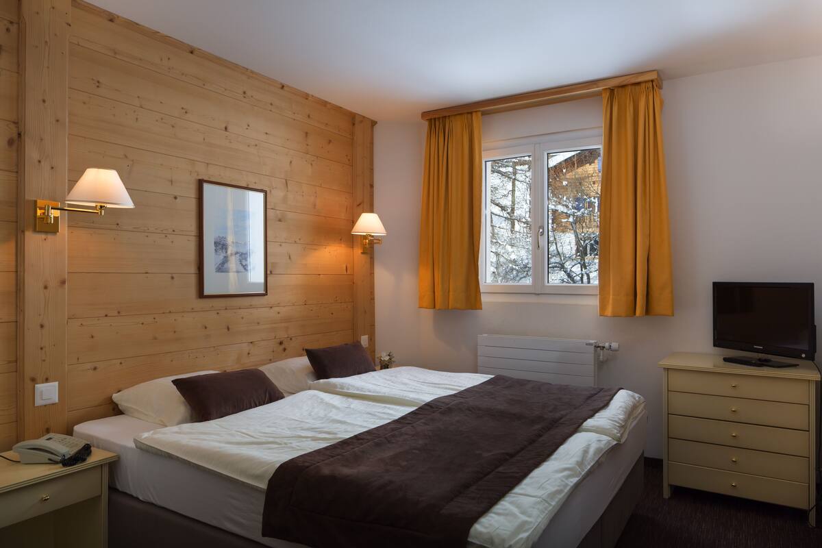 Une chambre avec un mur en bois, un lit avec une literie blanche et marron, deux lampes de chevet, un téléviseur posé sur une commode et une fenêtre avec des rideaux.