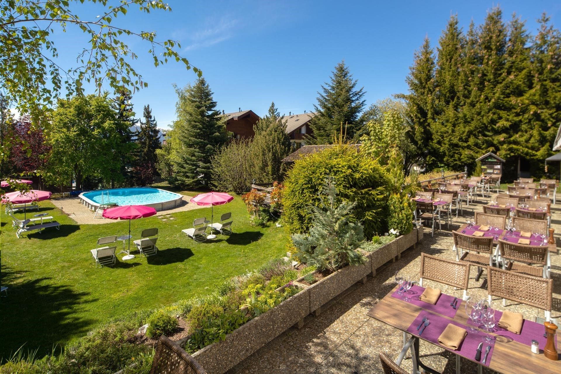 A pool area with lounge chairs and umbrellas, surrounded by trees and a dining area with tables and chairs.
