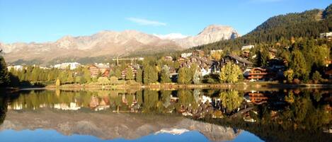 Un pueblo a orillas de un lago, con edificios reflejándose en el agua, rodeado de montañas.