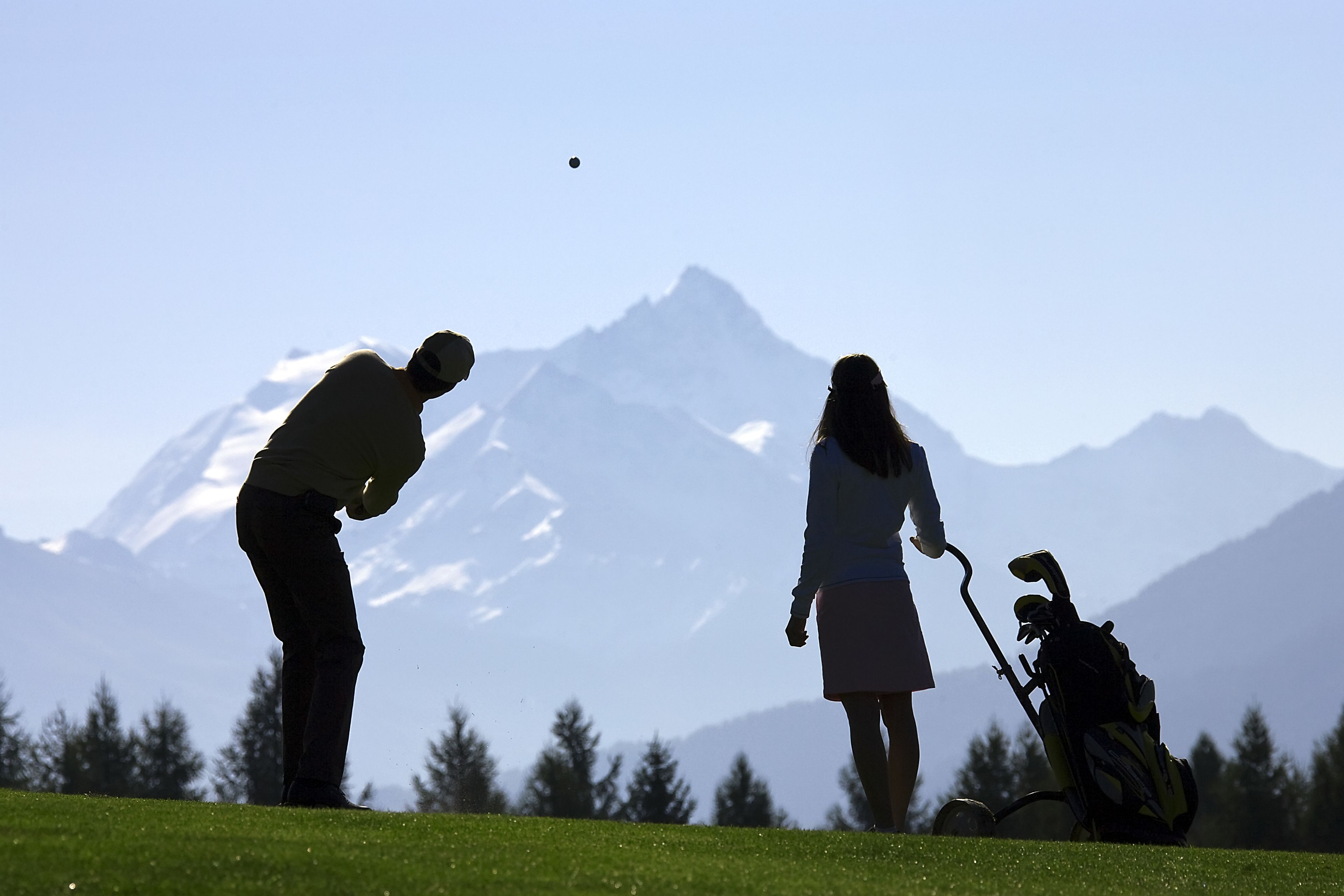 A silhouette of a person golfing with a golf bag and another person standing in a grassy area with mountains in the background.