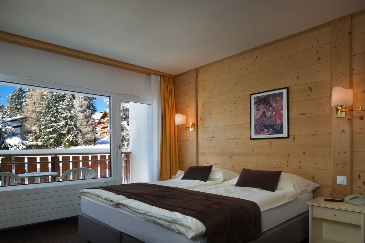 Une chambre d’hôtel avec un lit, une table de chevet, une lampe, un tableau au mur et une vue sur un paysage enneigé par la fenêtre.