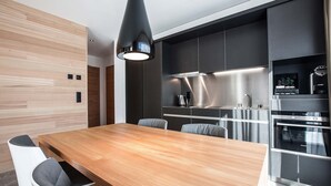 A modern kitchen with a wooden dining table and chairs, stainless steel appliances, and a black pendant light. - Hotel Belvedere (Scuol)