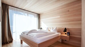 A modern bedroom with a wooden bed, a bedside table with a lamp, and sheer curtains. - Hotel Belvedere (Scuol)