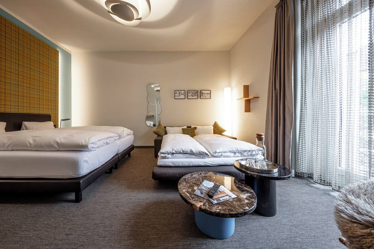 A hotel room with two beds, a round coffee table, a lamp, and a window with curtains.