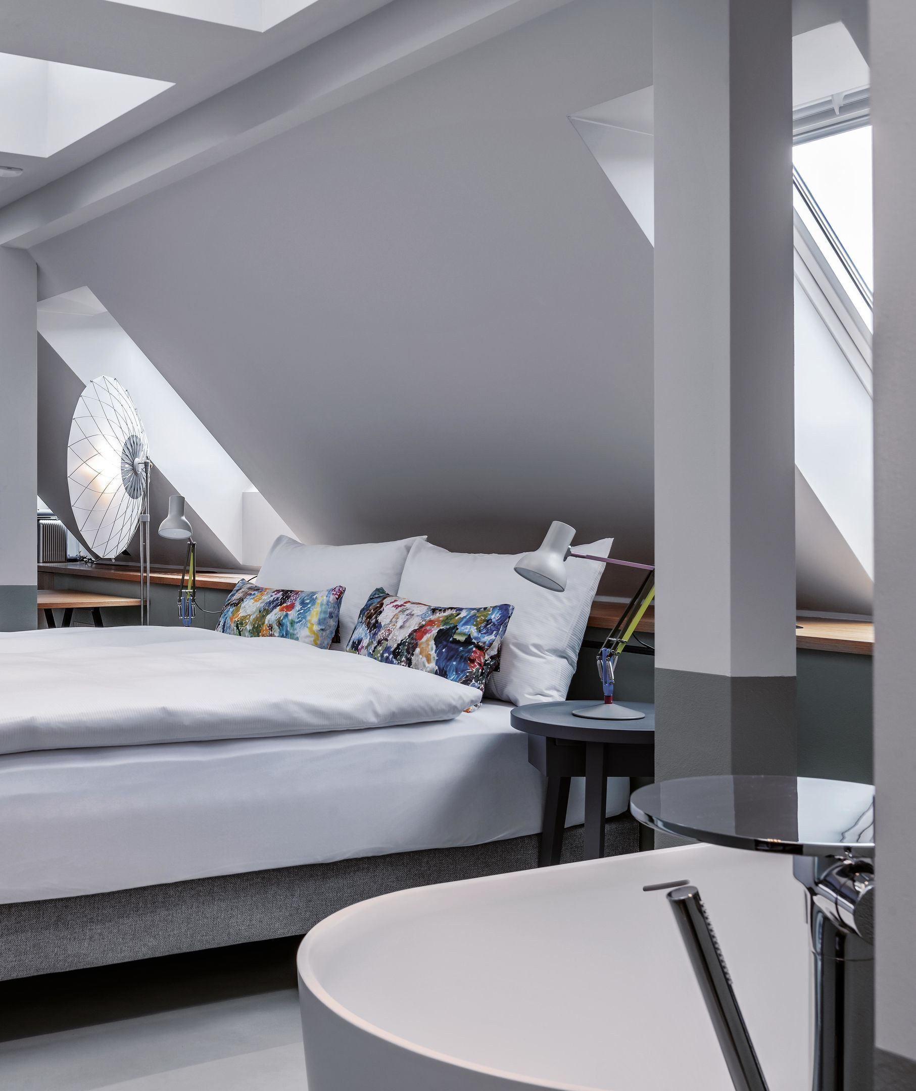 Un dormitorio moderno con un tragaluz, una cama con almohadones coloridos, una mesita de noche y un espejo redondo.